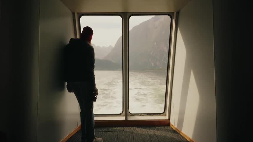 A silhouetted traveler with a camera looks out the back window of a boat at the majestic mountain fjords and the water's wake