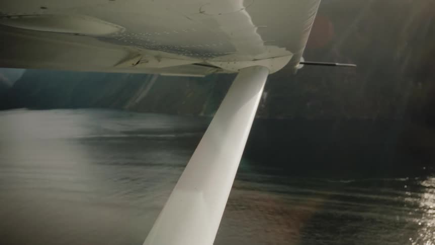 Underside of a small plane's wing as it flies over a majestic misty fjord with sun flare