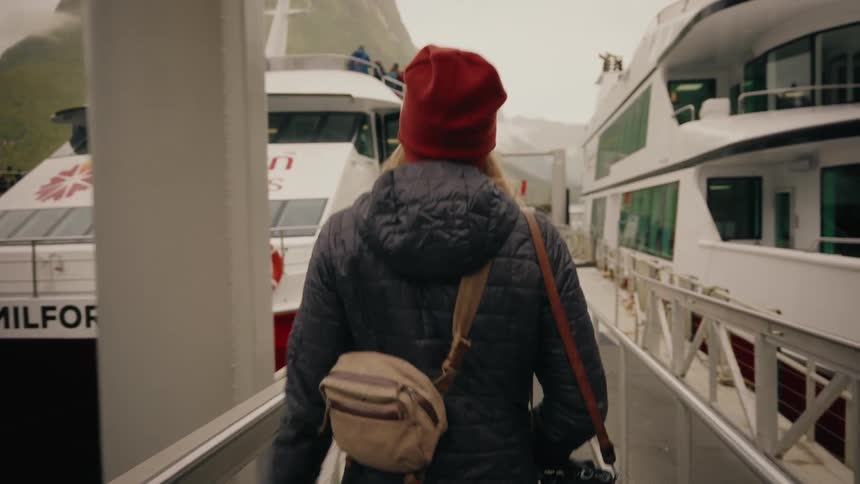 A traveler in a red beanie and puffy jacket walks down a gangway to board a tour boat at Milford Sound