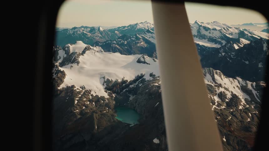 An aerial view from an airplane window shows a vast, snow-capped mountain range with a stunning turquoise glacial lake below