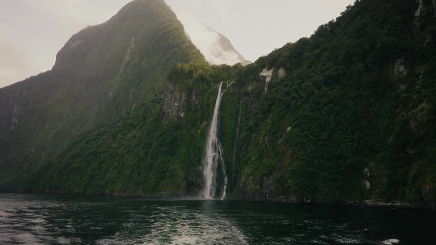 A majestic waterfall cascades down a steep, jungle-covered mountain into a fjord on a cloudy day