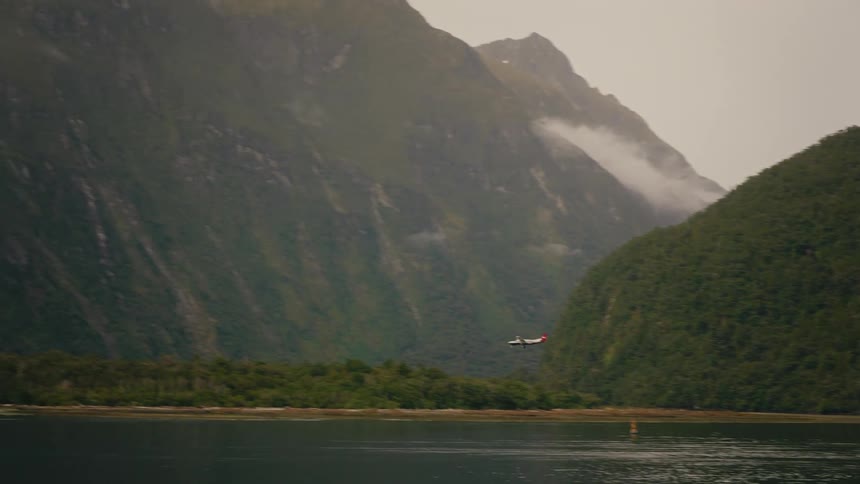 A small propeller plane flies low over the water through a dramatic mountain fjord