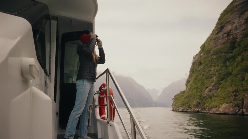 A female tourist with a camera emerges onto the deck of a boat to admire the breathtaking fjord scenery on a windy day