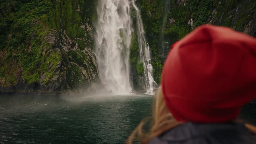 Over the shoulder shot of a woman in a red beanie watching a powerful waterfall cascade down a lush green cliff