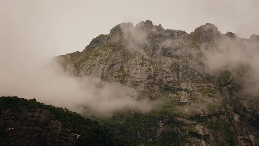 A towering, rugged mountain peak is shrouded in a thick layer of rolling fog on a cloudy day