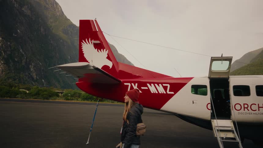 A young woman in a red beanie walks in slow motion across the tarmac of a remote mountain airfield after arriving on a small plane