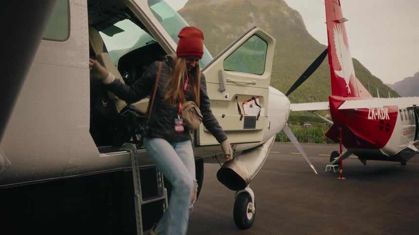 A young woman excitedly disembarks from a small plane and walks across the tarmac in a beautiful mountain setting