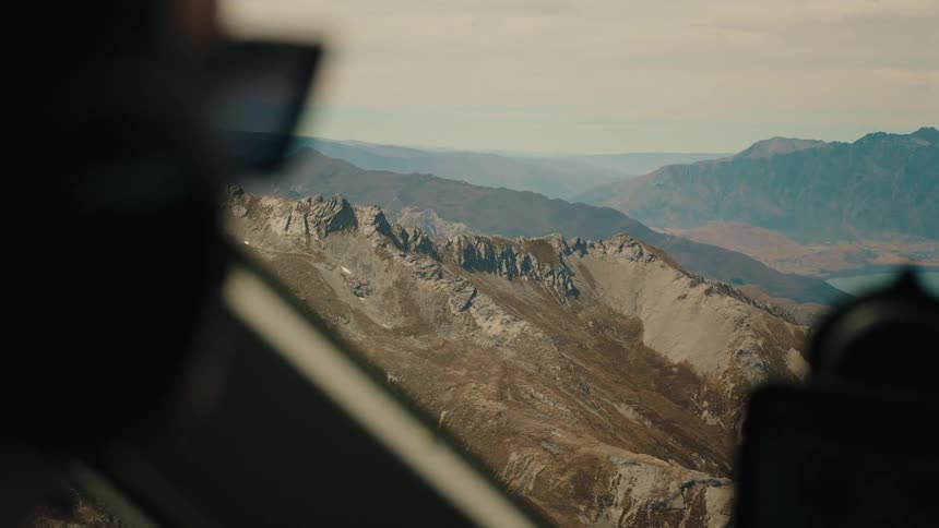 An over-the-shoulder perspective from a helicopter reveals a stunning, rugged mountain range and a distant lake