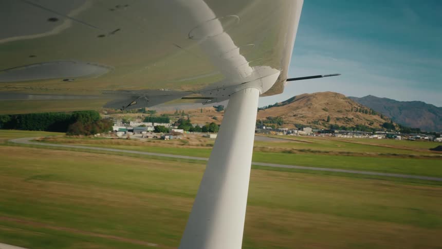 View from under the wing of a small airplane as it takes off from a runway surrounded by green fields and mountains