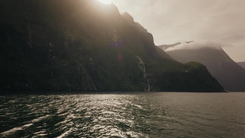 A cinematic shot from a boat revealing majestic green mountains and waterfalls lining a dramatic fjord as the sun peeks over the peaks