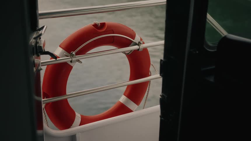 A red lifebuoy hangs on the railing of a sailing boat, with water visible in the background