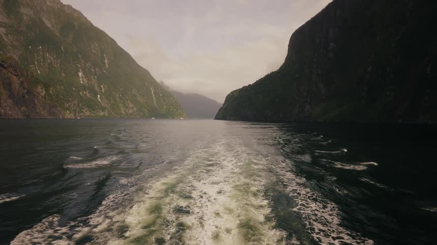 A boat's wake trails behind as it sails away through a majestic and misty fjord in New Zealand