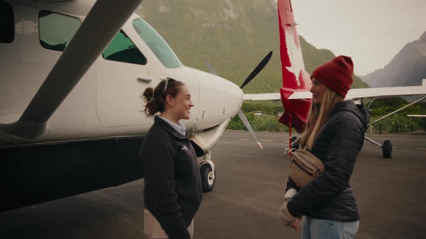 A female pilot and a tourist have a friendly conversation on the tarmac next to a small plane, with scenic mountains in the background