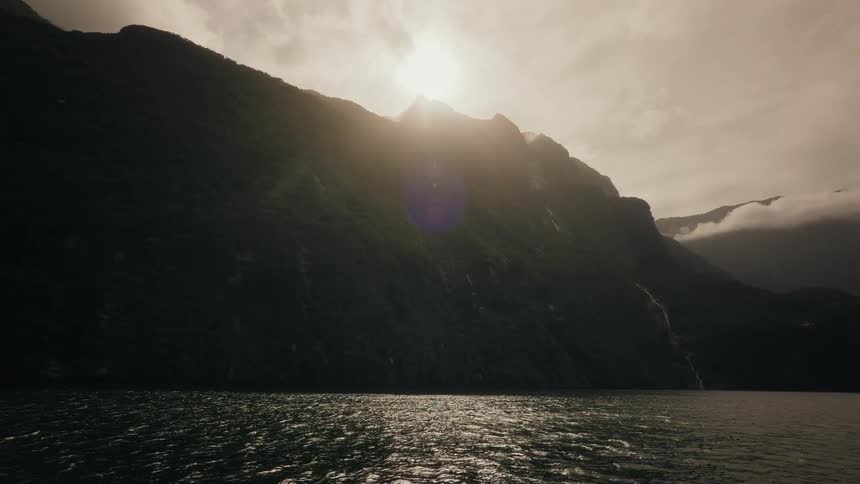 Sunlight crests over a silhouetted mountain range, casting a dramatic glare over the rippling waters of a majestic fjord