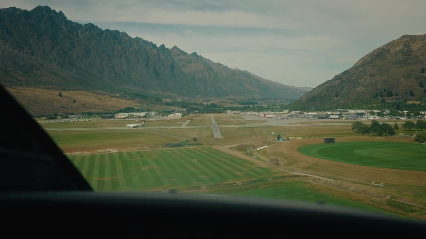 An airplane makes its final landing approach over green fields towards a runway in a mountain valley