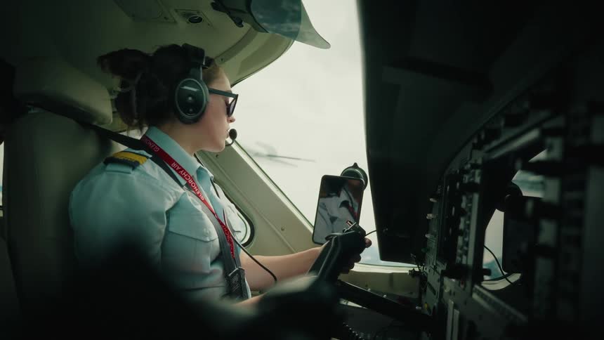 A woman pilot in uniform speaks into her headset and waves while navigating from the cockpit of a plane