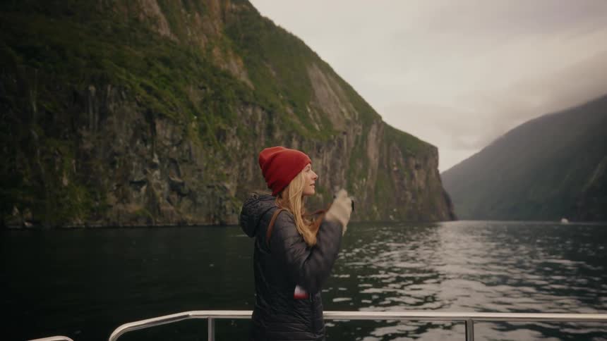 An adventurous young woman in a red beanie takes photos of a dramatic fjord on a scenic boat cruise