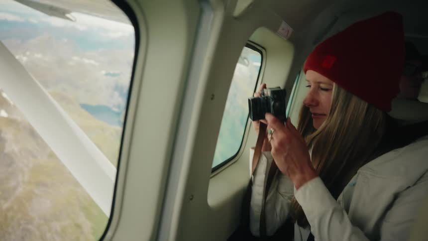 An adventurous woman in a red beanie photographs the stunning mountain scenery from the window of a small airplane