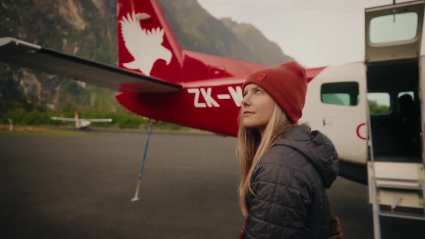 An adventurous young woman in a red beanie waits on a remote mountain airfield to board a small propeller plane