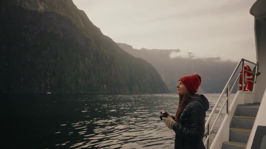 A female photographer in a red beanie stands on a boat deck, capturing the breathtaking views of a misty fjord