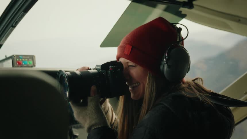 A happy female photographer in a red beanie smiles while taking photos from inside the cockpit of a small airplane
