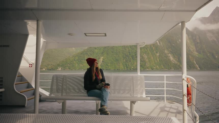A young woman with a camera sits on a tour boat bench, enjoying the scenic mountain views of a fjord