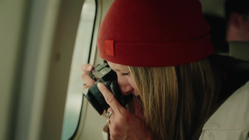 A happy young woman in a red beanie smiles while looking out the window of a small airplane before taking photos of the landscape below