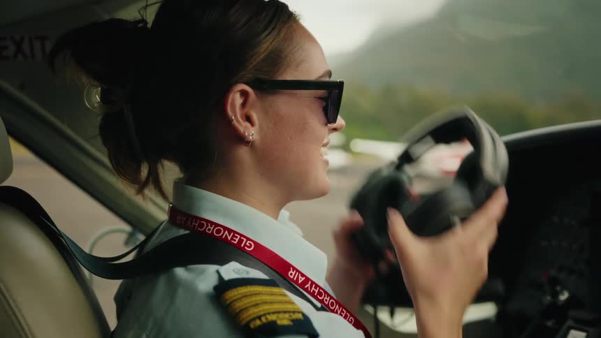 A confident female pilot prepares for takeoff in the cockpit, putting on her sunglasses and headset with a smile