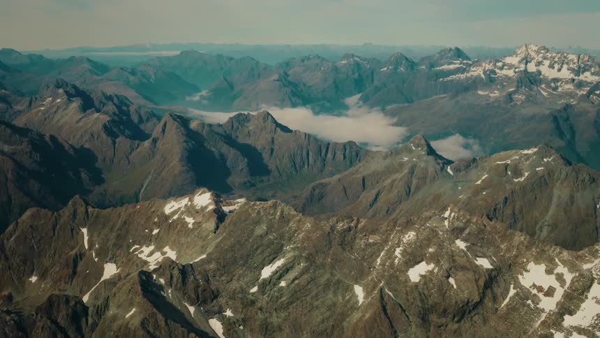 An epic aerial view slowly pushes over a majestic, snow-dusted mountain range with clouds nestled in the deep valleys