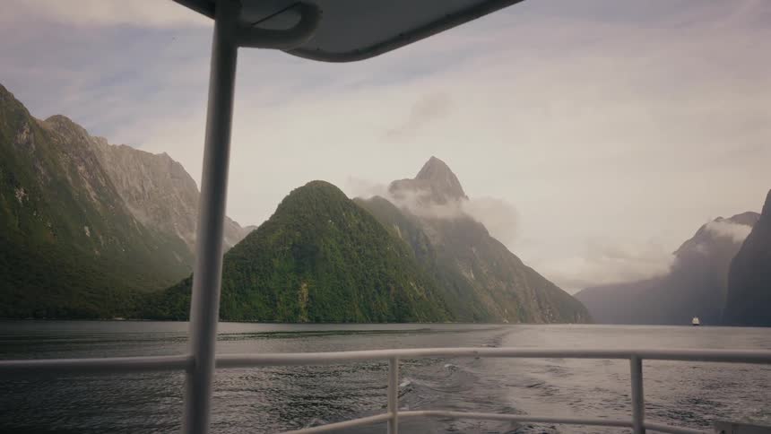 A scenic view from the stern of a tour boat reveals the majestic, cloud-covered mountains of a fjord as it sails away