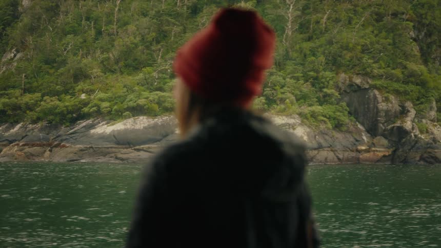 A cinematic shot of a tree-covered, rocky shoreline seen from the water, with the silhouette of a person in a red beanie in the foreground