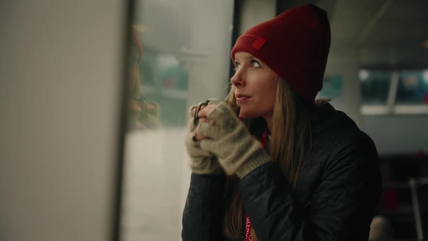 A woman wearing a red beanie and warm gloves thoughtfully sips a hot beverage while looking out a window during her travels