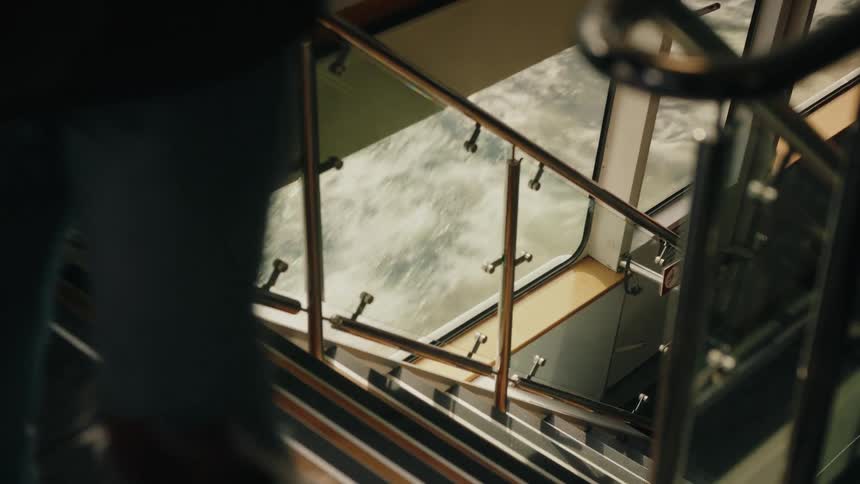 A traveler with a camera walks up the stairs of a moving ferry past a window showing the choppy sea