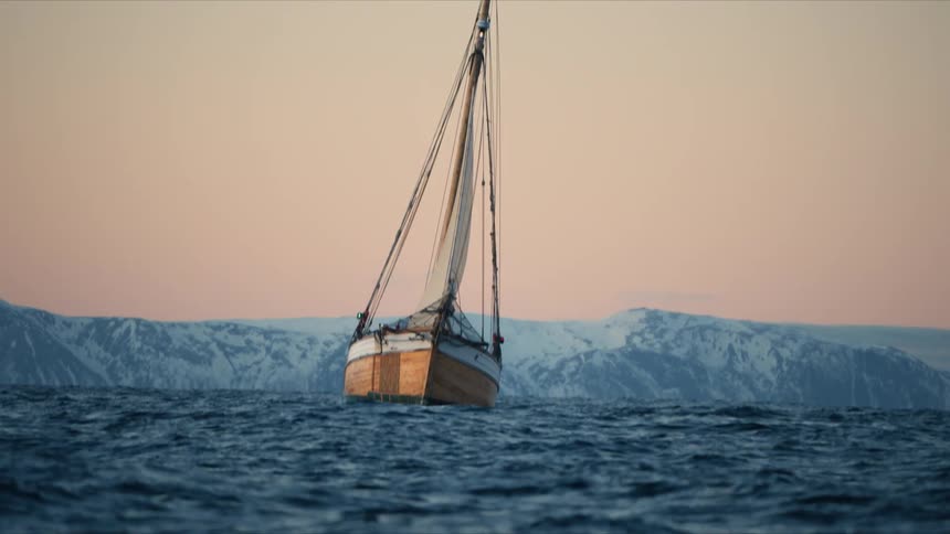 A traditional wooden sailboat sails across the open sea with majestic snow-covered mountains in the distance at sunset