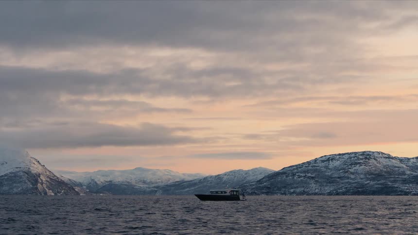 A lone boat drifts peacefully on a vast fjord at sunset, framed by majestic snow-capped mountains under a serene cloudy sky
