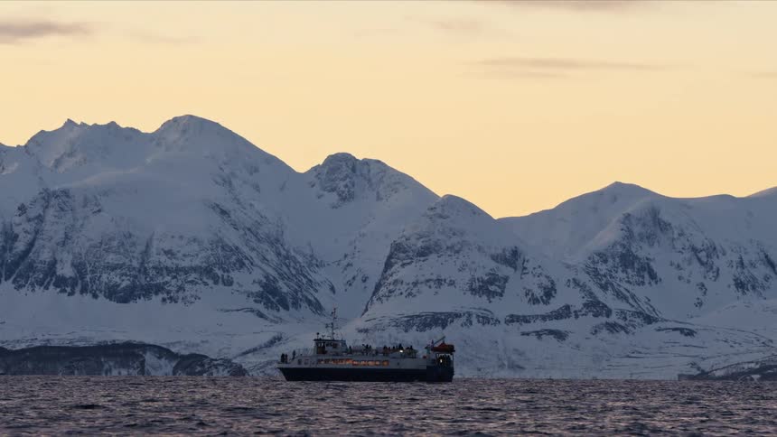 A passenger ferry glides across a tranquil fjord at dusk with a stunning backdrop of snow-covered mountains