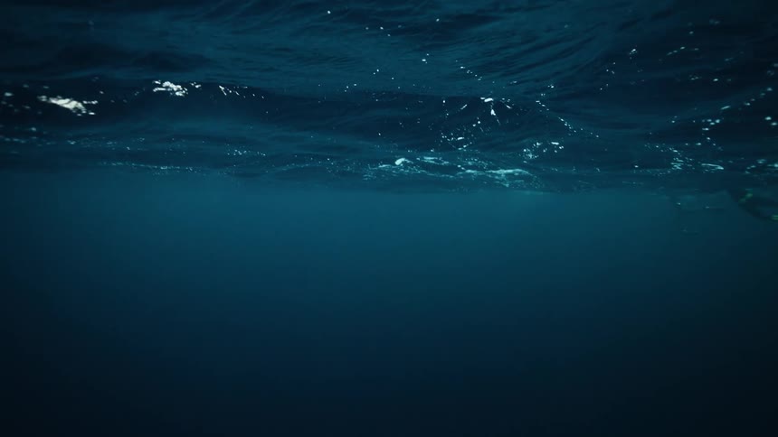 A slow-motion underwater view looking up at the deep blue ocean surface as it gently ripples
