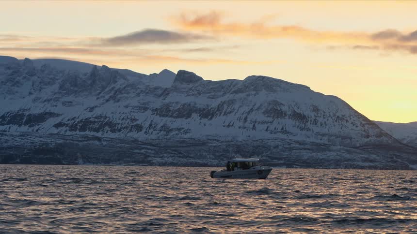 A scenic view of a boat floating on a tranquil sea against a backdrop of majestic, snow-covered mountains at sunset