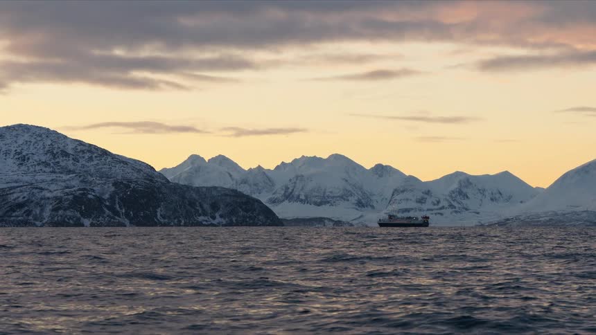A passenger ferry peacefully sails across a calm sea, framed by a stunning backdrop of snow-covered mountains under a serene sunset sky