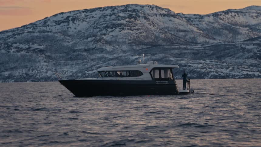 A lone figure stands on a boat as it gently rocks on the water in front of majestic, snow-capped mountains at sunset