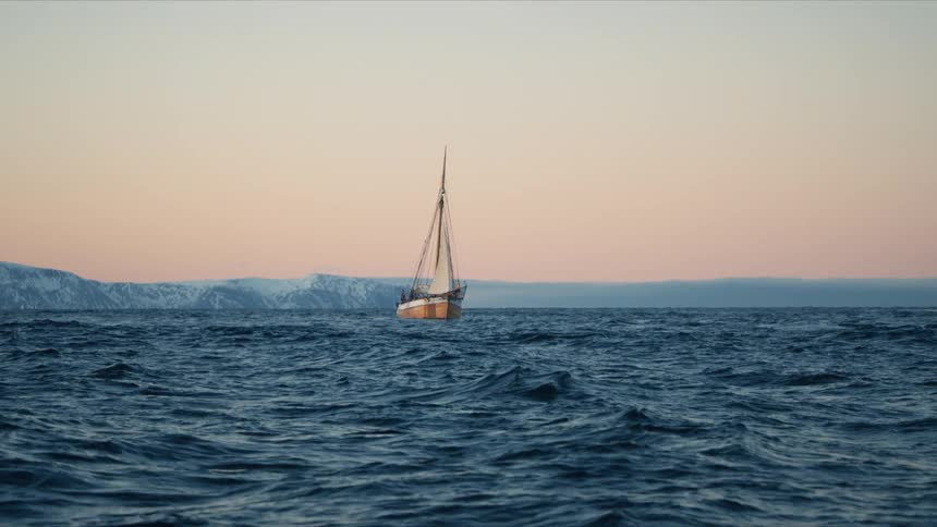 A solitary sailboat traverses the restless arctic sea at dusk, with snow-covered mountains on the horizon