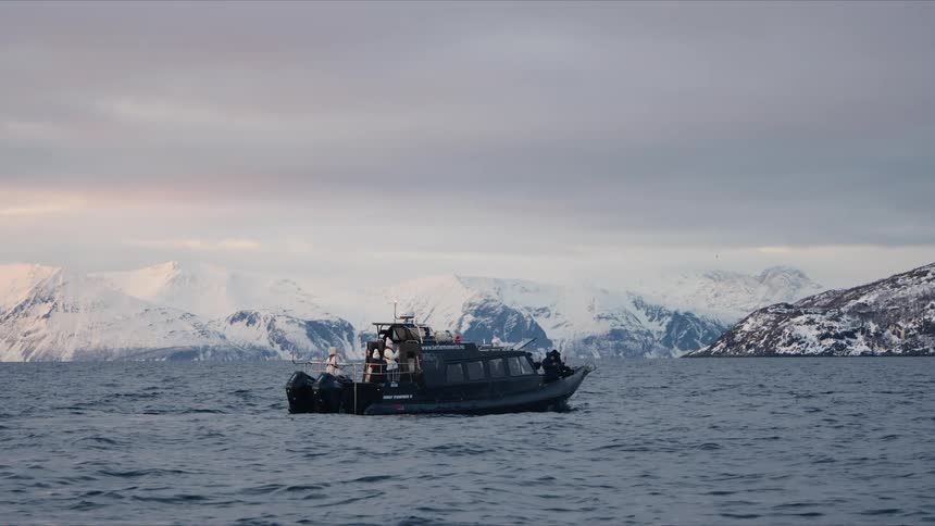 A tour boat filled with passengers gently floats on the calm waters of a fjord, with a breathtaking landscape of snowy mountains in the background