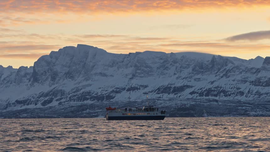 A tour boat with passengers enjoys the scenic view of a majestic snow-covered mountain range from the water during a vibrant winter sunset