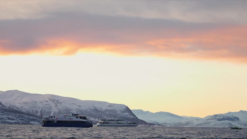 Two passenger boats sail across a serene fjord in Norway during a winter sunset, against a backdrop of snowy mountains