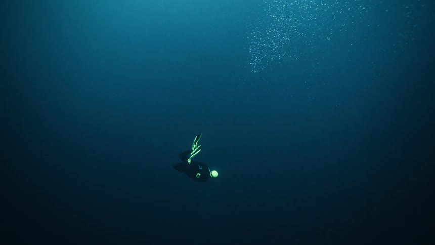 A lone scuba diver descends into the dark blue abyss of the ocean, turning on a flashlight to explore the depths