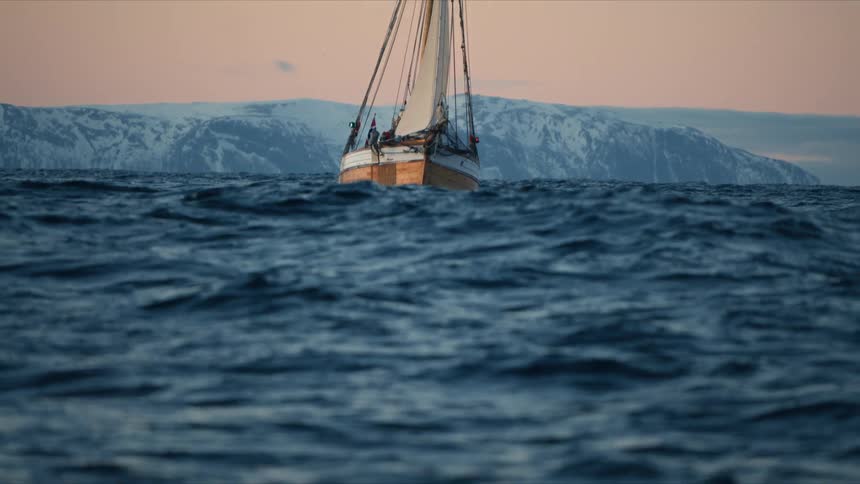 A classic wooden sailboat sails on a choppy arctic sea towards snowy mountains at dusk as the focus shifts to the waves