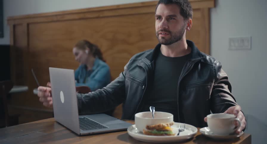 A businessman has a video conference call on his laptop while enjoying lunch at a modern cafe