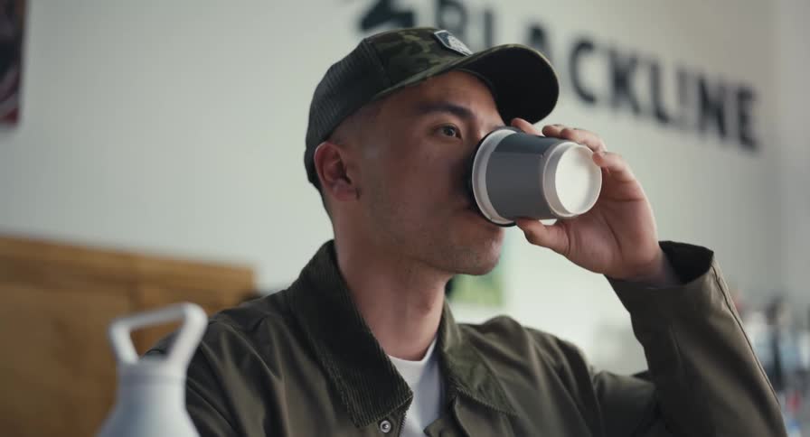 A young man wearing a camouflage cap sips coffee from a disposable cup while engaged in a conversation