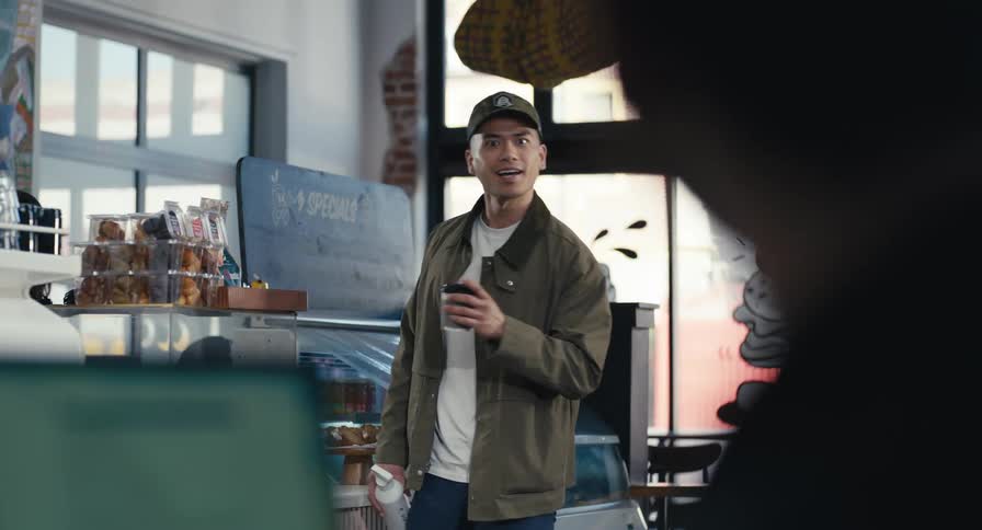 A stylish man in a coffee shop gets his drink from the counter, turns, and is surprised before smiling warmly