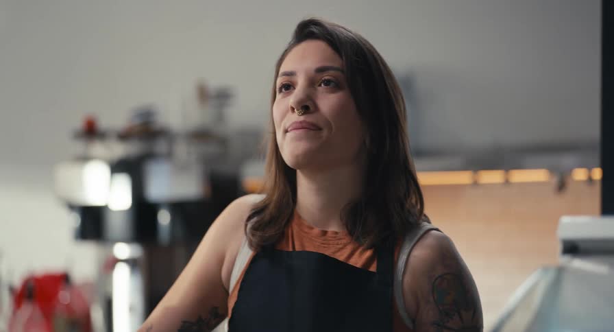 A friendly female barista with tattoos smiles as she takes an order from a customer in a coffee shop